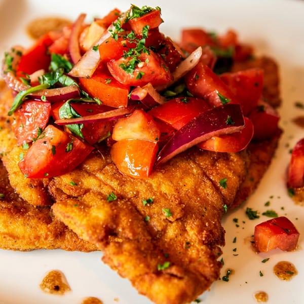 Golden fried cutlet topped with fresh diced tomatoes, red onions, and herbs on a white plate.