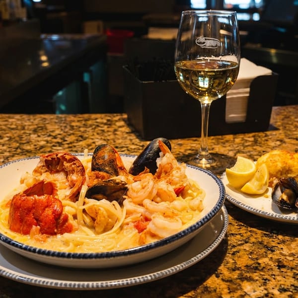 Seafood pasta with lobster, shrimp, and mussels, a glass of white wine, and a side dish on a bar counter.